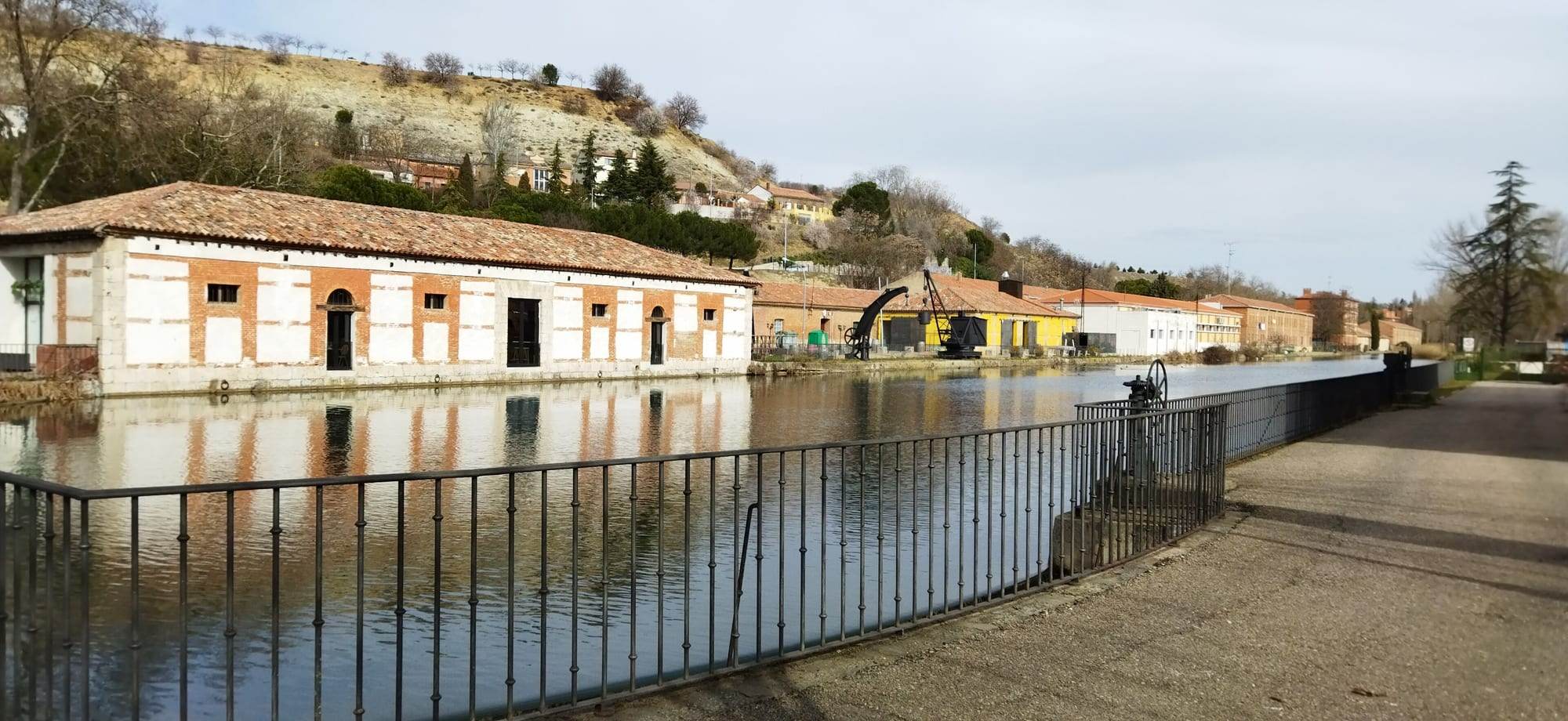 The Dársena, the basin where the Canal de Castilla ends in Valladolid: a long whitewashed warehouse banded with red brick under a terracotta tile roof is mirrored in the still water, two old black iron cranes stand on the far quay in front of brick and yellow buildings, a wrought-iron railing and a sluice wheel line the towpath in the foreground, and a scrubby hillside dotted with houses rises behind under a pale sky